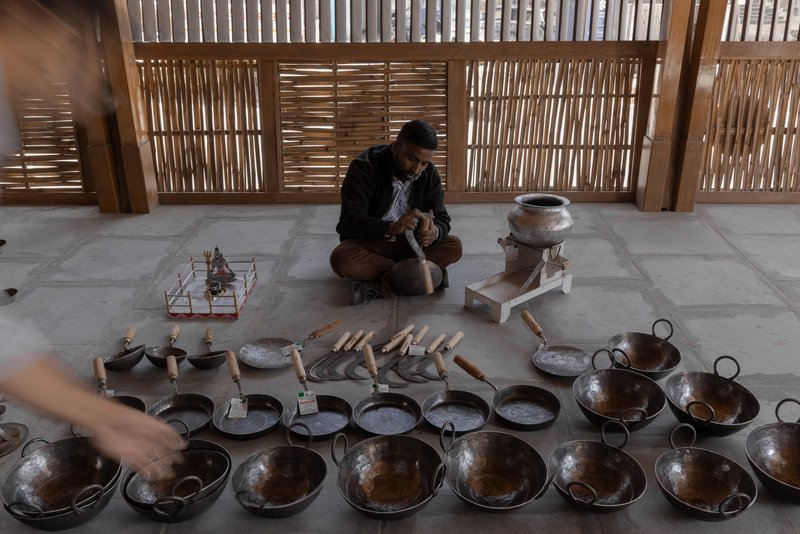 Seated artisan working among rows of metal cookware and handtools beneath slatted timber screen wall