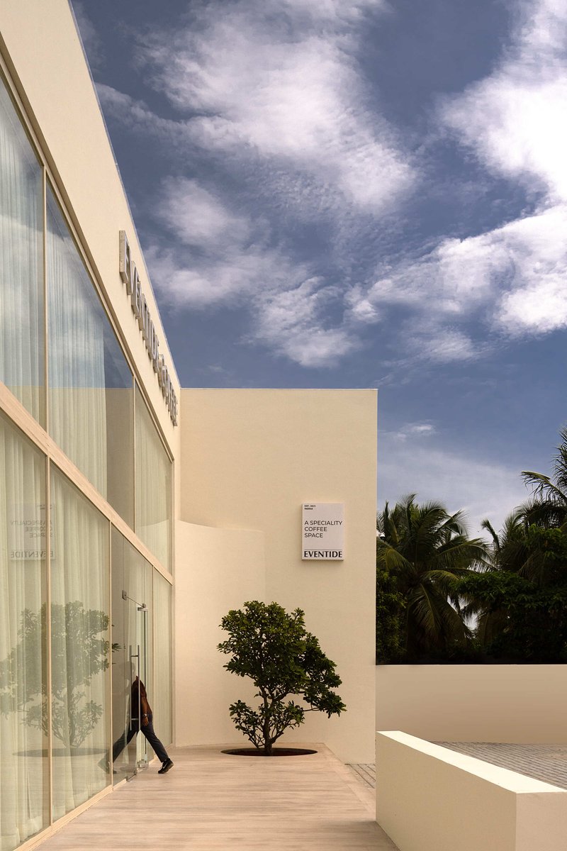 Exterior walkway with translucent glass facade and single tree planted in paved terrace under cloudy sky