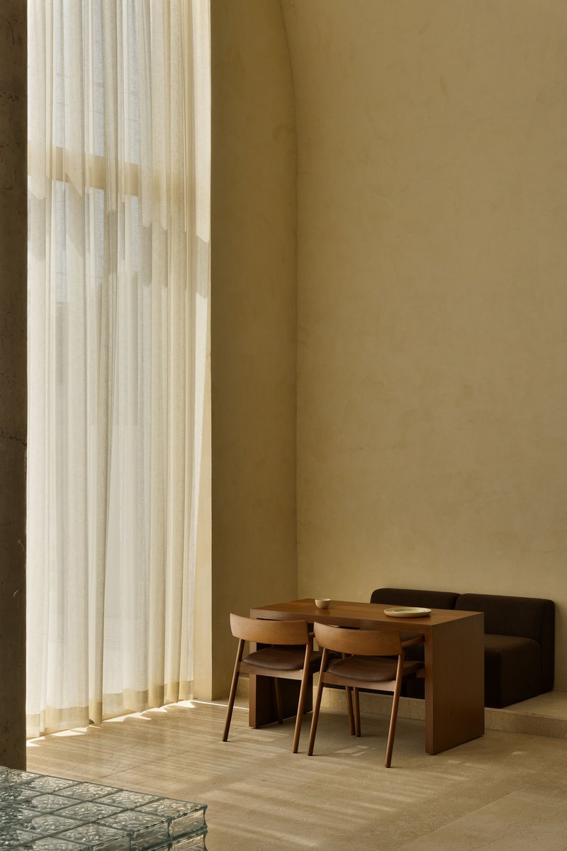 Interior dining area with timber table and chairs beside floor-to-ceiling sheer curtains in warm daylight