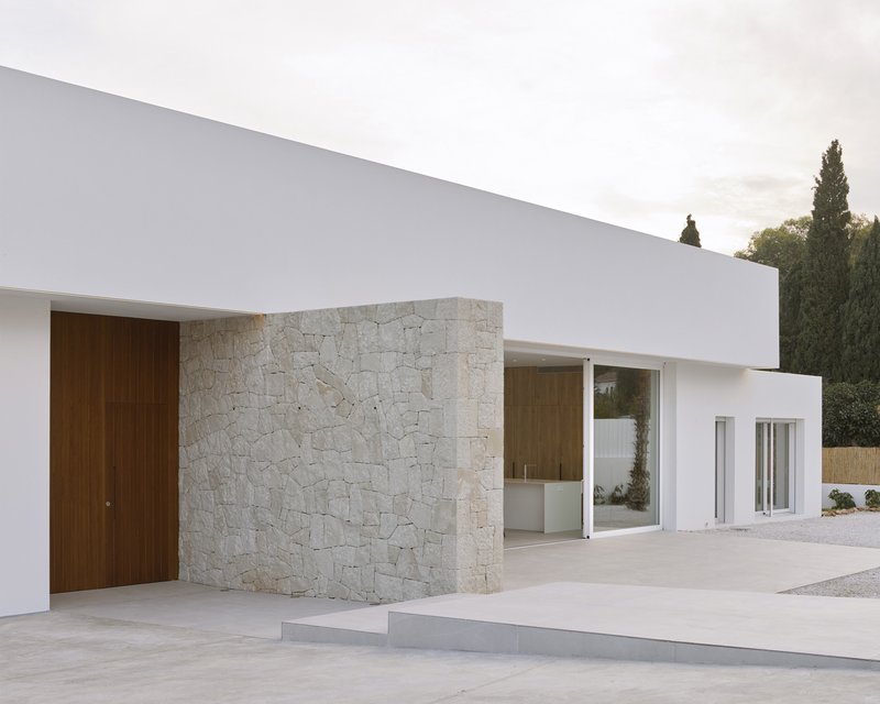 Facade composition of white stucco volumes and limestone cladding with cypress trees visible beyond