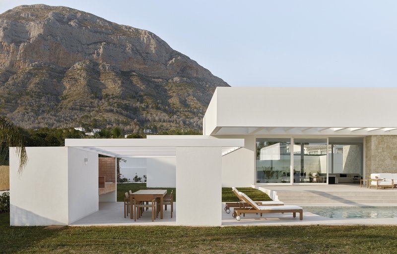 Garden view showing white stucco volumes with a covered dining pavilion and pool deck against a rocky hillside