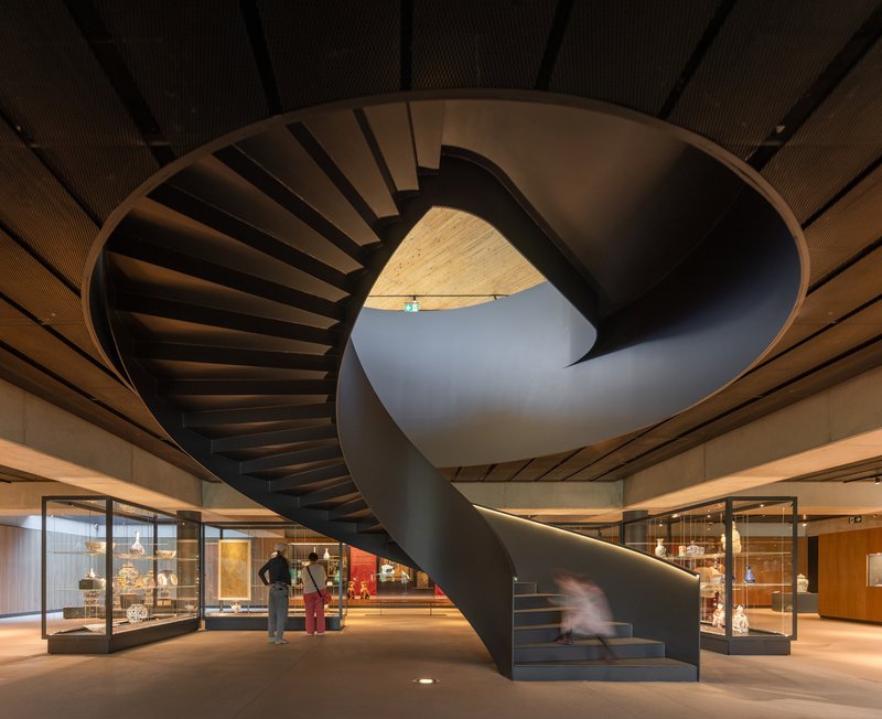 Spiral staircase in dark steel rising through the gallery space with timber-slatted ceiling above