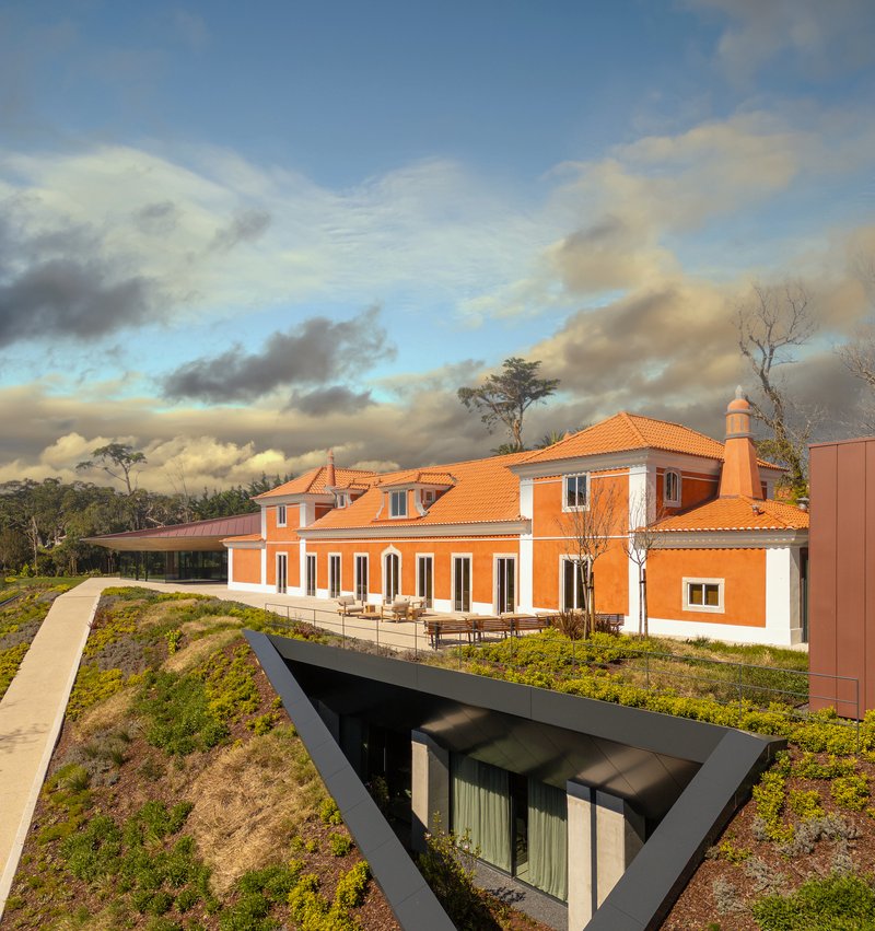 Orange stucco villa with terra-cotta roof beside green roof planes under afternoon clouds