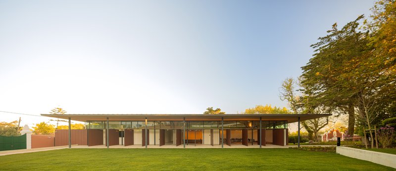 Glass and steel pavilion with overhanging roof on a lawn at golden hour with trees flanking