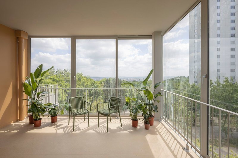 Enclosed balcony with glazed corner walls, potted plants and two chairs overlooking green treetops