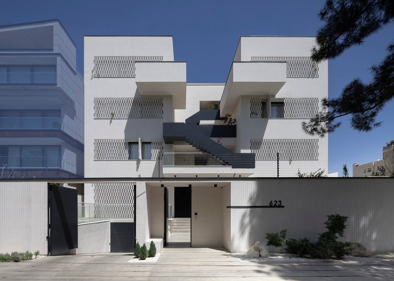 Street facade with stacked balconies and perforated white screens behind a concrete wall with planted entry court