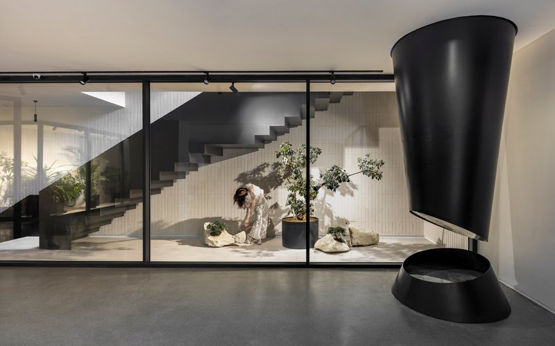 Interior view through glazed partition showing cantilevered black staircase and person tending bonsai plants