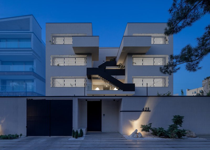 Street facade at dusk with white corrugated metal fence and illuminated perforated balcony screens