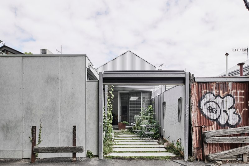 Narrow laneway entrance framed by concrete panels and corrugated metal shed with weathered timber fence