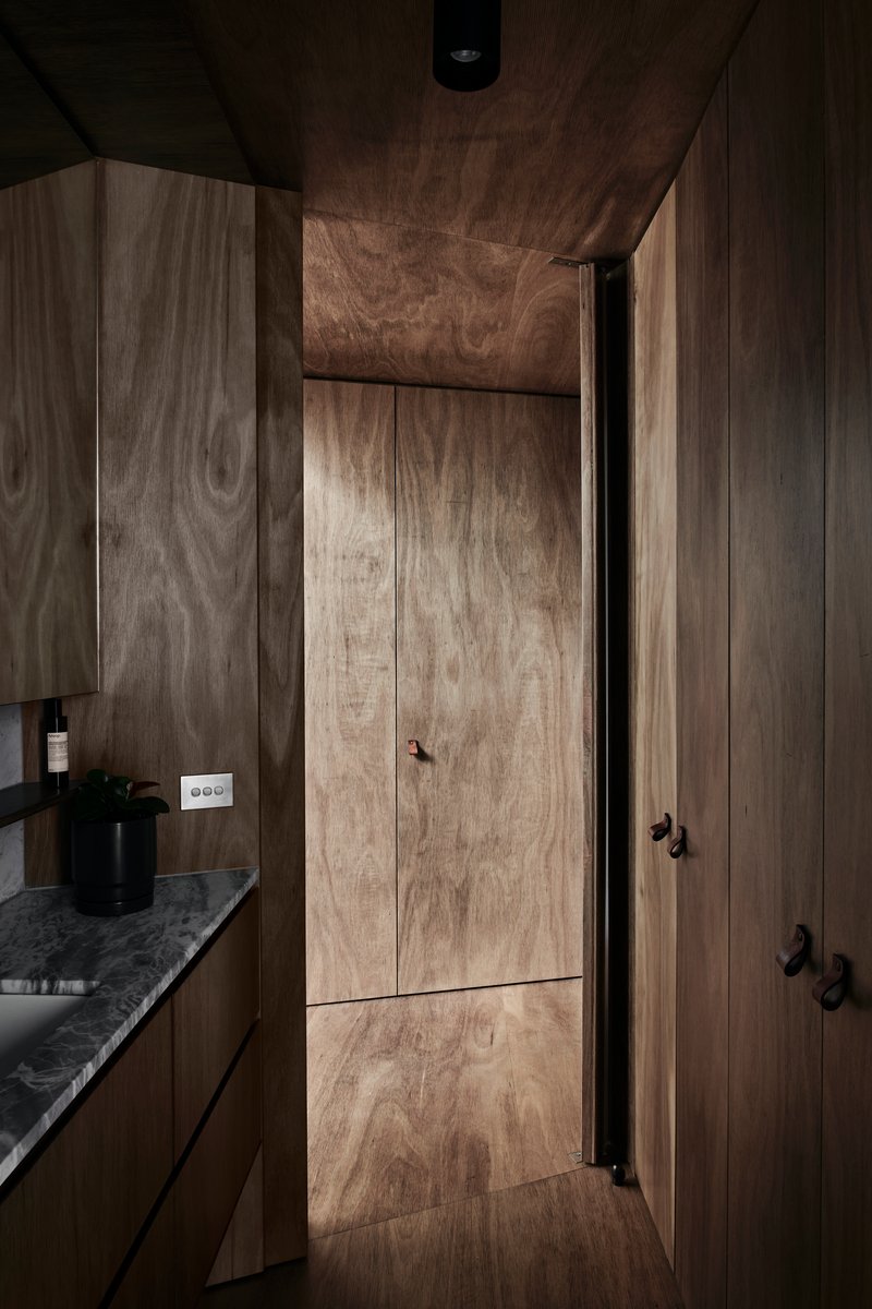 Narrow corridor clad entirely in timber veneer with flush doors and a stone countertop glimpsed beyond