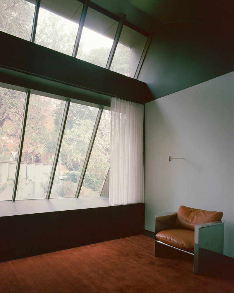 Bedroom with angled windows and timber beams overlooking garden vegetation through sheer curtains