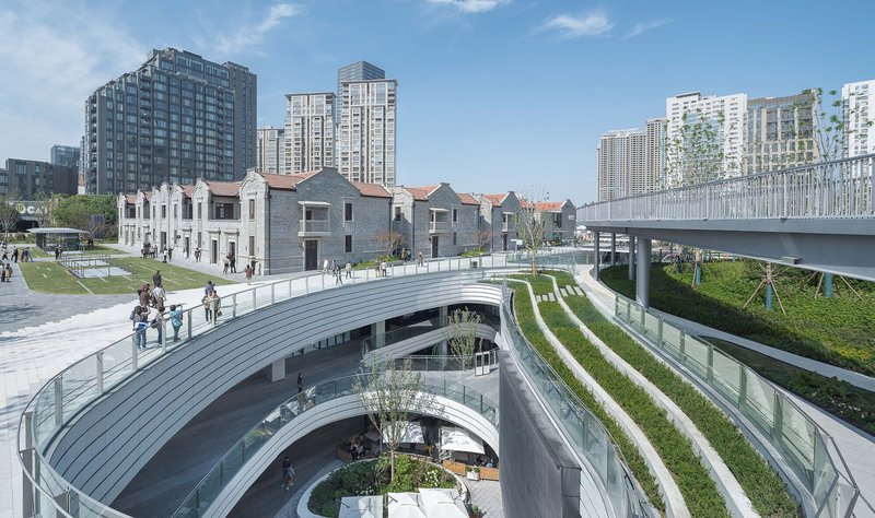 Elliptical sunken plaza with striped grass terraces and perimeter glass balustrades framing preserved brick buildings