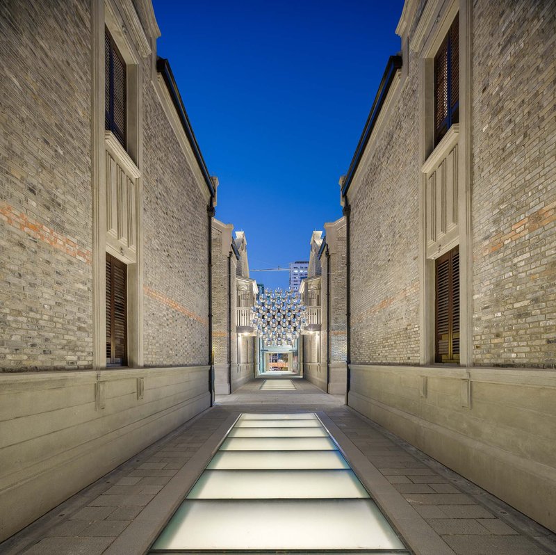 Narrow lane with illuminated glass floor panels between grey brick walls at dusk