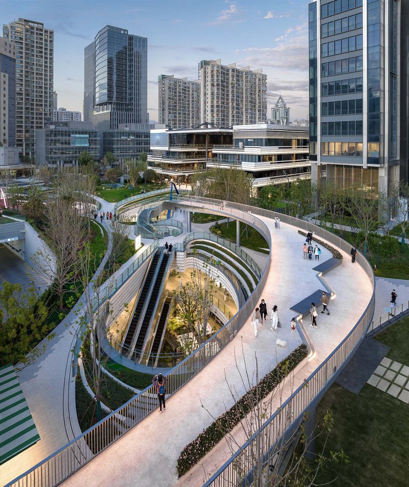 Spiraling pedestrian ramps descending around a sunken courtyard with trees at dusk