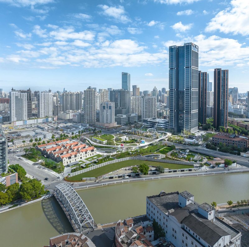 Aerial view of a landscaped park with curving pathways along a river under partly cloudy skies