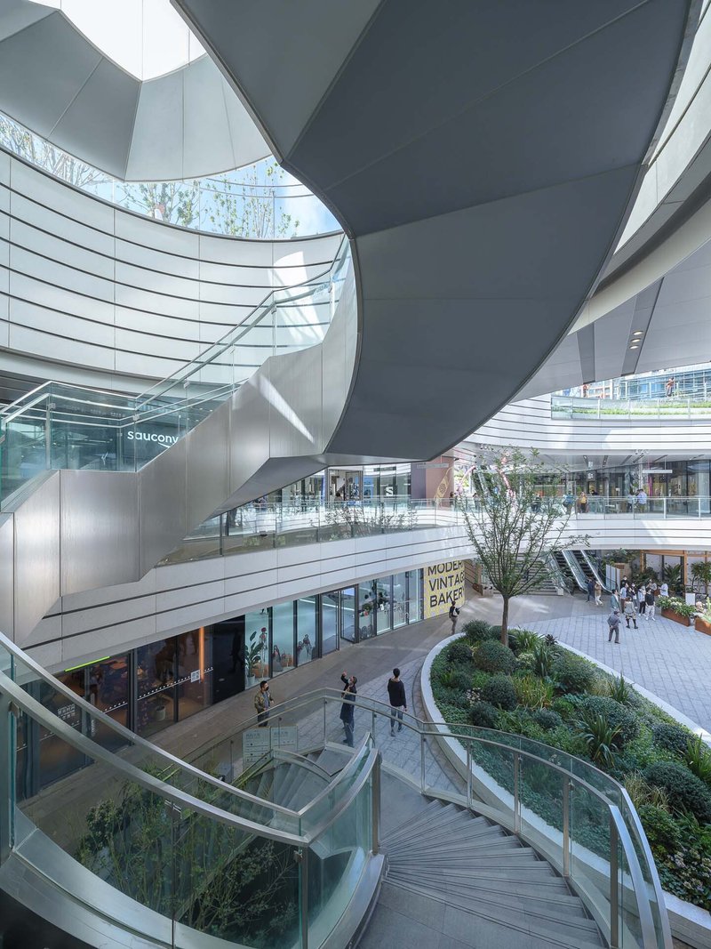 Multi-level atrium with curved white balconies, glass railings, planted terraces and shoppers on escalators below