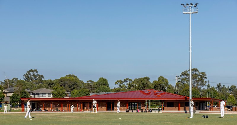 Long brick pavilion with red metal roof and deep eaves beside a lawn bowling green under clear sky