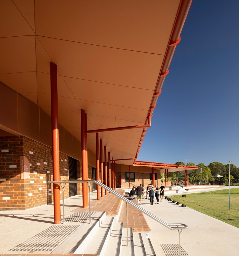 Covered walkway with steel columns and cream ceiling panels alongside variegated brick facade in shadow