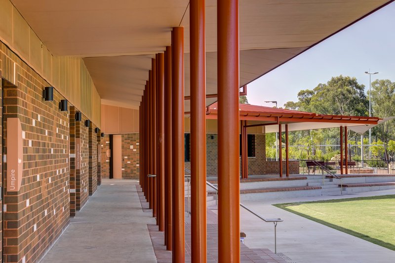 Covered walkway with reddish brown cylindrical columns along a multicolored brick wall under afternoon light