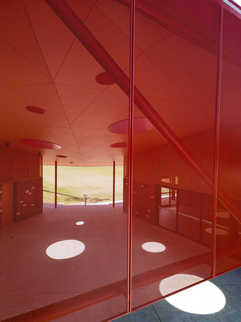 Reflections of circular ceiling apertures and columns on red perforated metal panels in afternoon sunlight