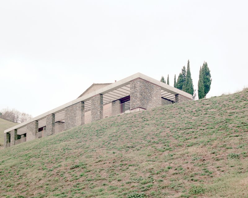 Stone-clad residence emerging from a grassy hillside with cypress trees under an overcast sky