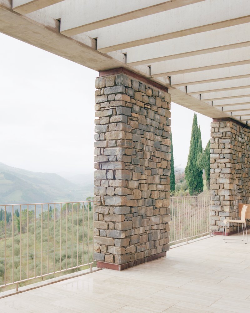 Covered terrace with handlaid stone columns and timber ceiling joists overlooking distant valley and cypress trees