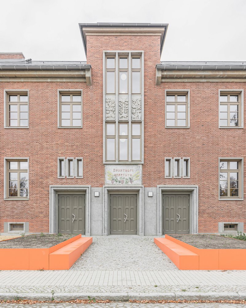 Red brick facade with tall glazed bay and orange benches flanking the entrance under overcast sky