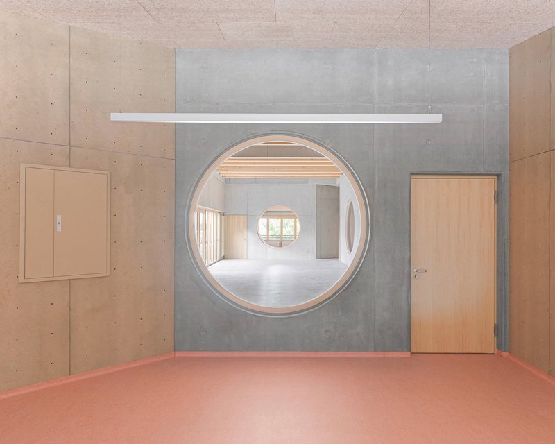 Circular portal in a concrete wall revealing a sequence of rooms with pink flooring and timber ceilings
