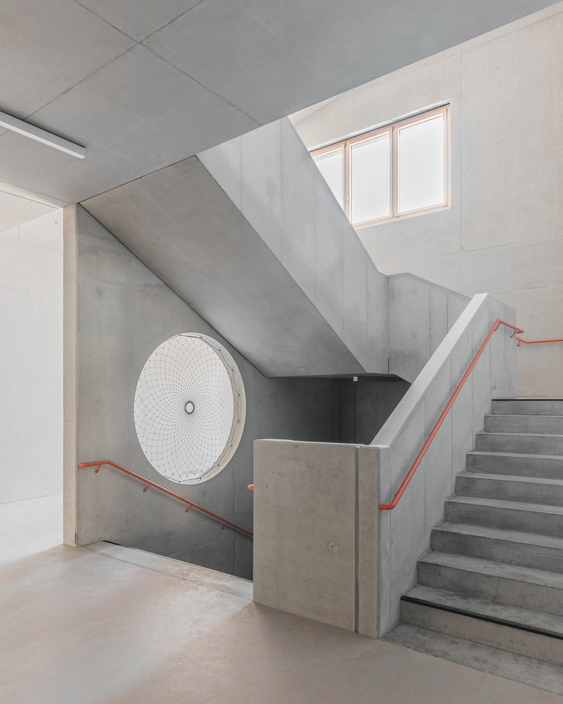Concrete stairwell with orange handrails ascending toward a skylight above a circular decorative grille
