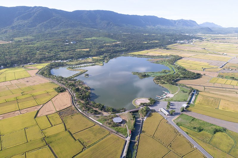 Aerial view of pavilions and curved roofs beside a reservoir surrounded by rice fields and mountains