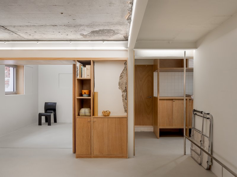 Light wood cabinetry and shelving units dividing white-walled rooms beneath an exposed concrete ceiling
