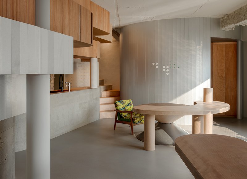 View across the curved timber table toward the sunlit grey wall and wooden staircase rising to the mezzanine