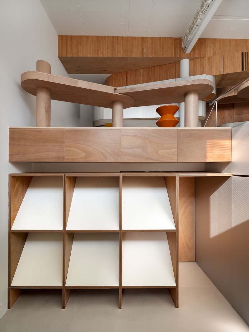 Interior view of plywood shelving unit beneath a curved timber handrail and cylindrical columns in soft daylight