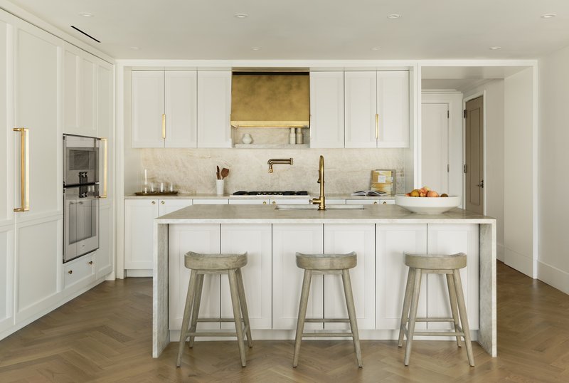 Kitchen island with three upholstered stools and brass range hood against white cabinetry