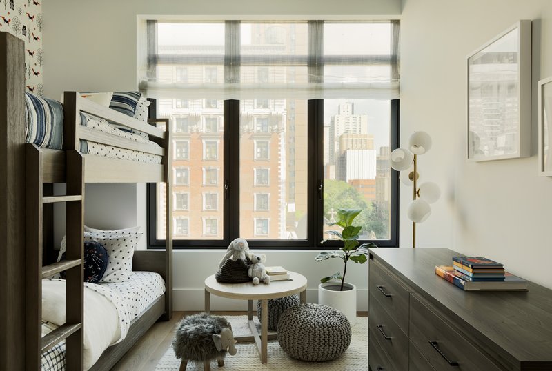 Bedroom with bunk bed and black-framed windows overlooking urban apartment buildings through sheer roller shades