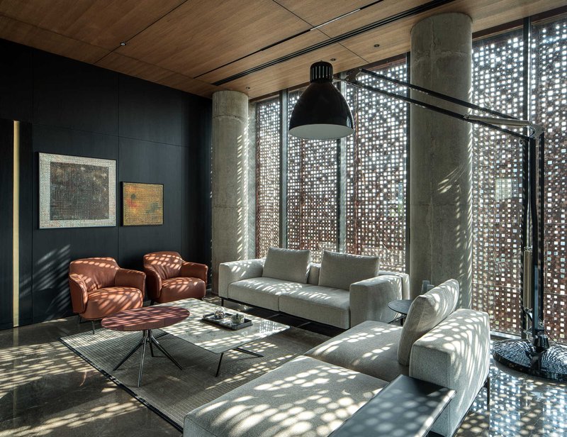 Double-height lounge with perforated metal facade, exposed concrete columns and dappled light patterns across timber ceiling