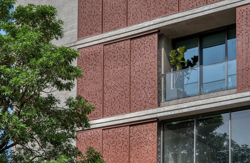 Perforated terracotta facade panels with concrete slab edges and glass balconies framed by green foliage