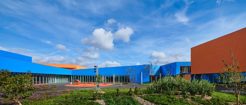 View across planted landscape toward the horizontal blue and orange volumes with glazed corridors in afternoon light