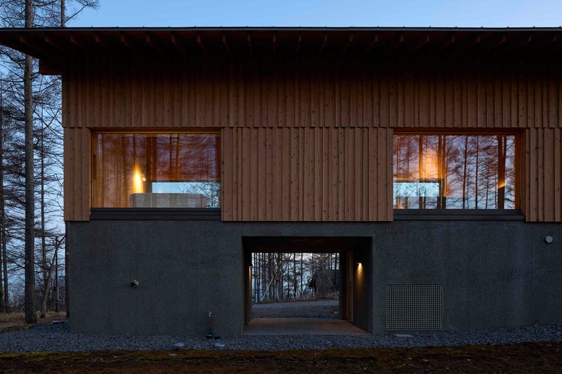 Two-story facade with vertical timber cladding over dark plaster base and covered passageway at dusk