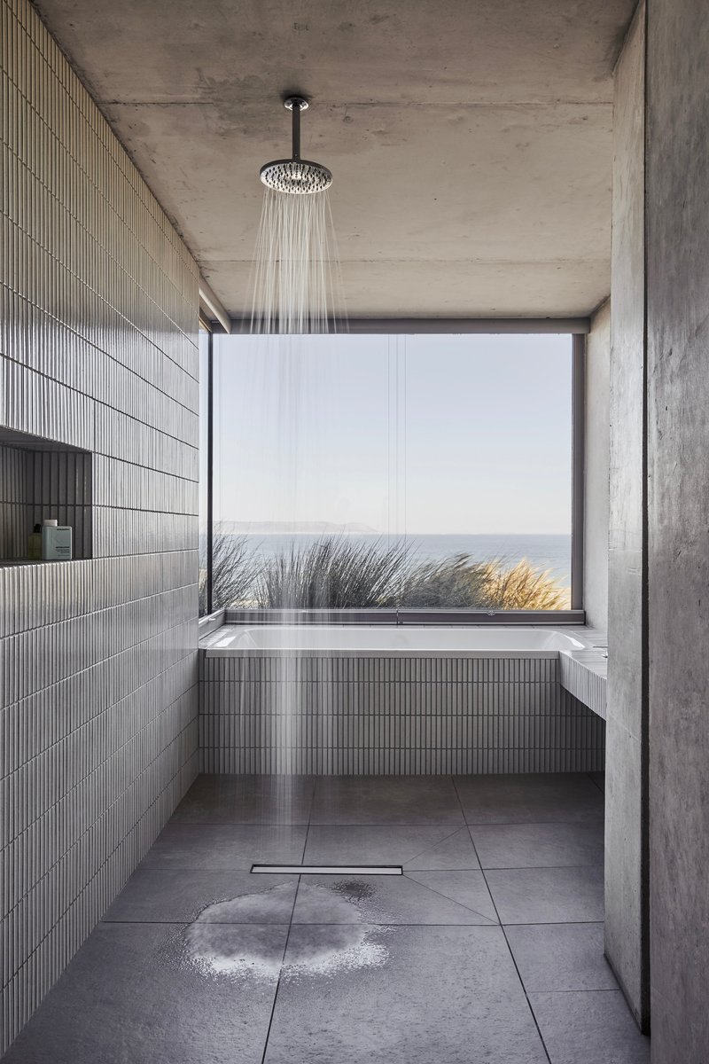 Concrete bathroom with rainfall shower, ribbed tile and picture window framing coastal grasses and ocean
