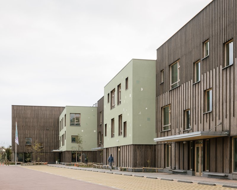 Street view showing the junction of pale green rendered volumes and vertical timber-clad blocks under overcast sky