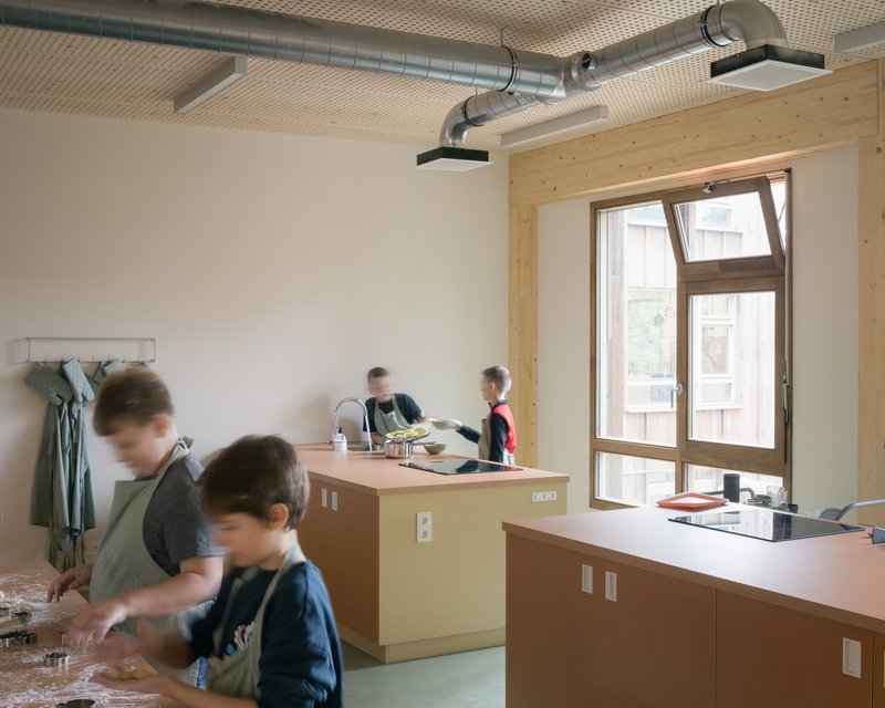 Kitchen workspace with exposed ventilation ducts and children gathered around countertops near a tall window
