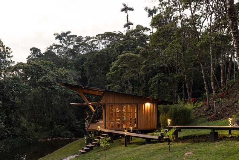Elevated timber pavilion on pilotis with folded roof and screened facade at dusk beside a pond