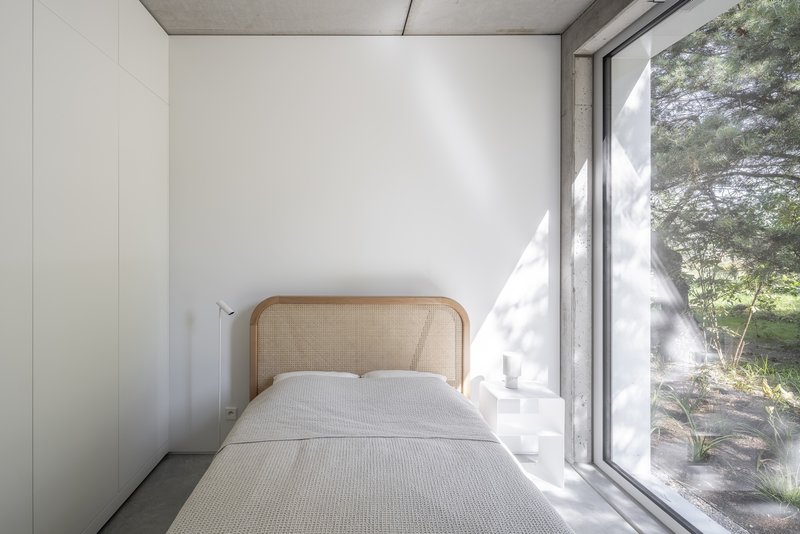 Bedroom with floor-to-ceiling glazing casting dappled sunlight across the white walls and concrete ceiling