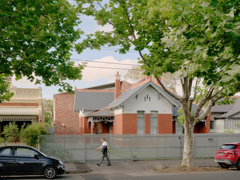 Street view of a gabled house with red brick base and grey painted timber under green tree canopy