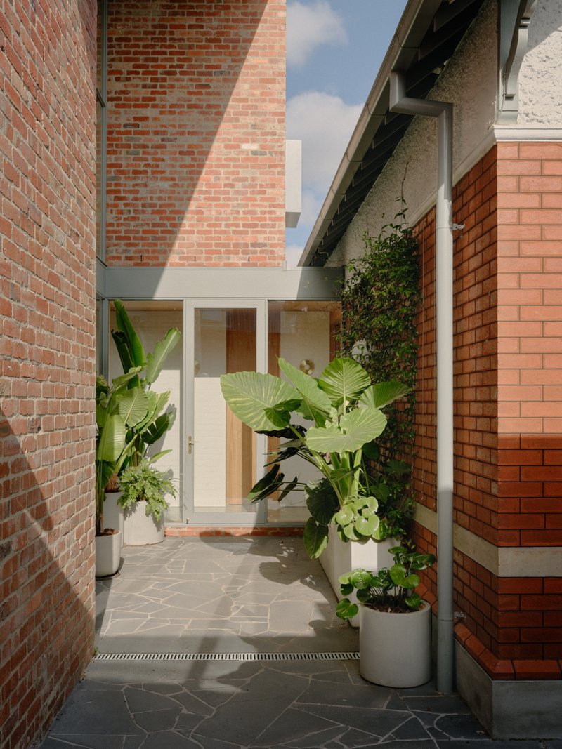 Narrow courtyard passage with red brick walls, potted banana plants and grey stone pavers in sunlight