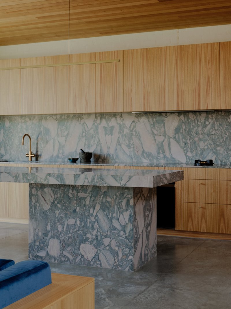 Kitchen island clad in pale green and white terrazzo with timber cabinetry under slatted ceiling