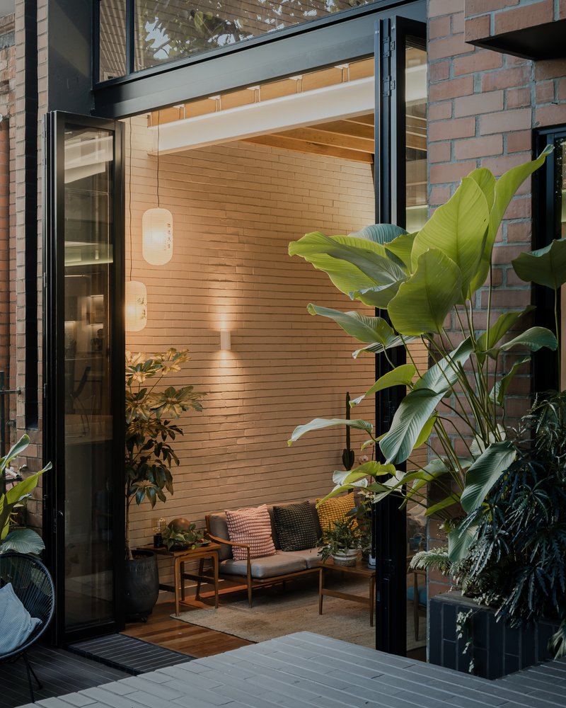 Courtyard view through black-framed sliding doors with timber cladding and tropical planting at dusk