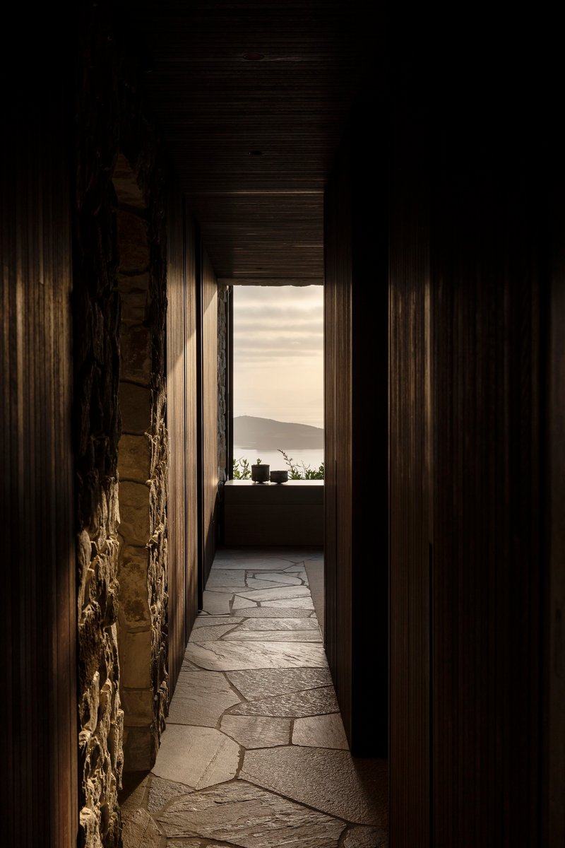 Narrow corridor with dark timber walls and irregular stone flooring leading to distant coastal view