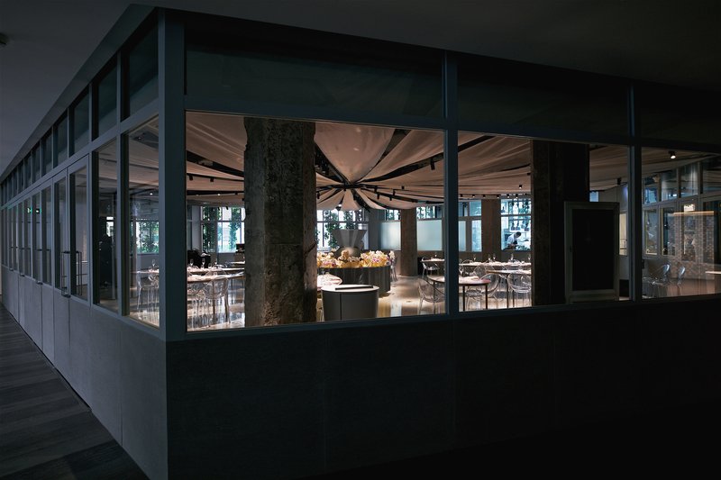 Exterior view through glazed facade showing the illuminated dining room with draped ceiling at dusk